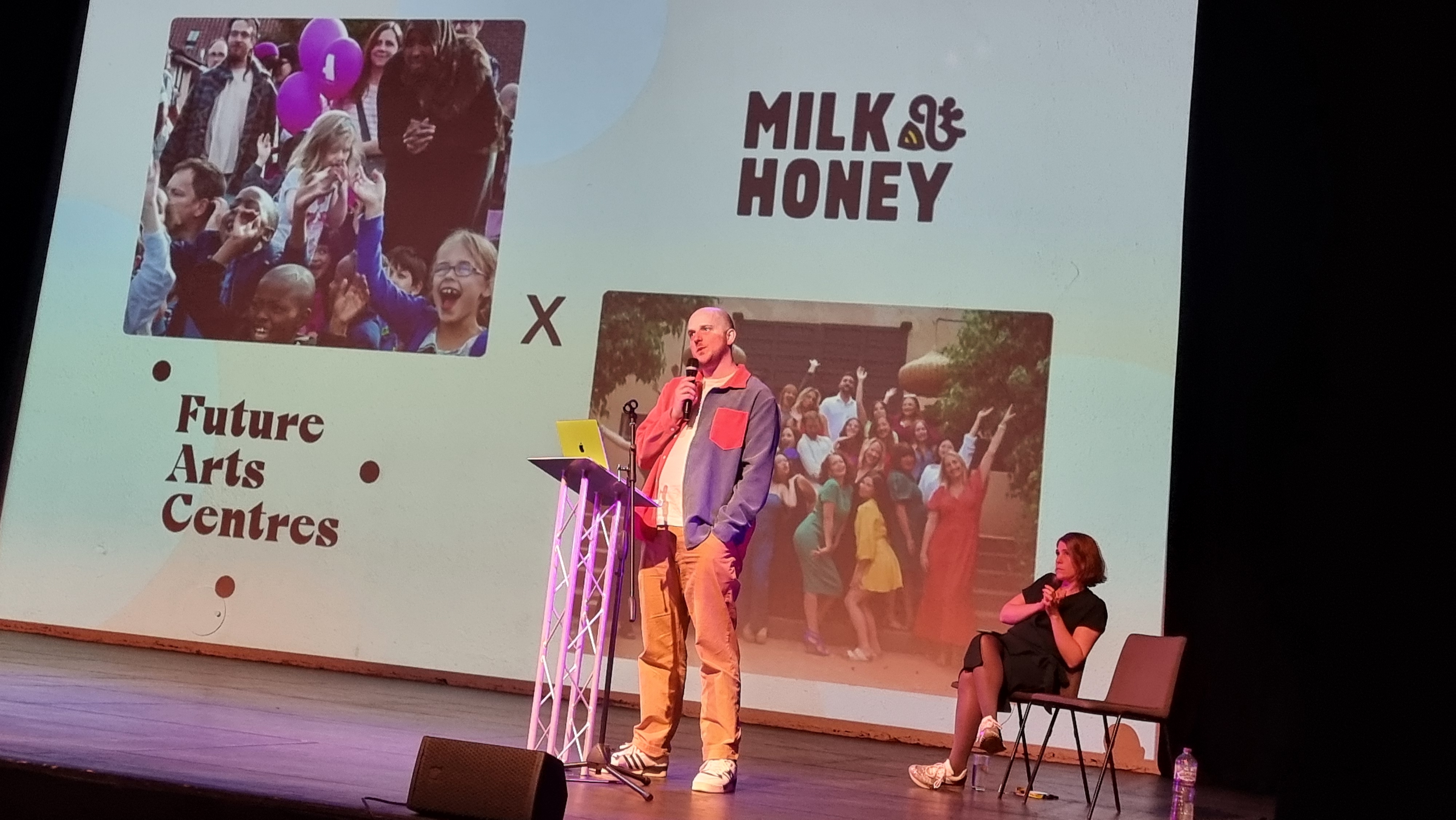 Two people on stage, one seated, and one stood at a metal lectern, presenting to an audience (out of shot). Behind them is a large projection screen, with two photos showing activity at arts centres; the Future Arts Centres logo; and the Milk & Honey logo