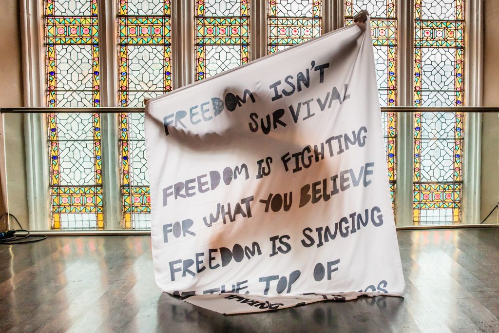 A person (hidden) holding up a large piece of material with text reading 'Freedom isn't survival, freedom is fighting for what you believe. Freedom is singing...' (rest of text not visible)