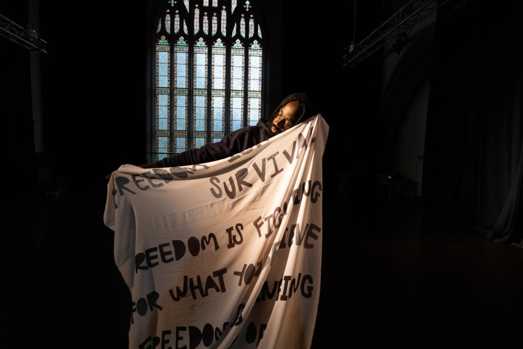A person (only their head and one arm are visible) holding up a large piece of material with text reading 'Freedom isn't survival, freedom is fighting for what you believe. Freedom is singing...' (rest of text not visible)