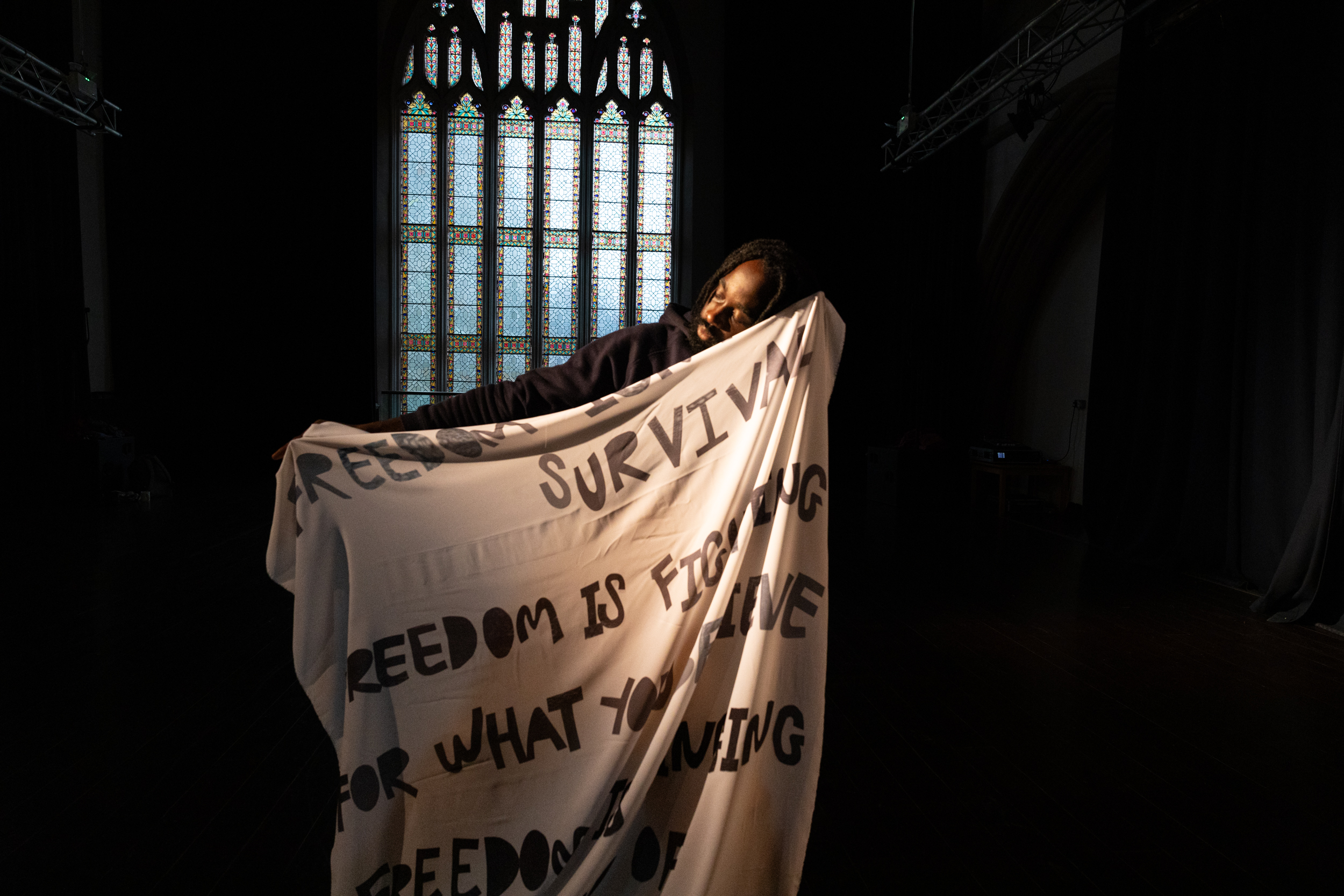 A person (only their head and one arm are visible) holding up a large piece of material with text reading 'Freedom isn't survival, freedom is fighting for what you believe. Freedom is singing...' (rest of text not visible)
